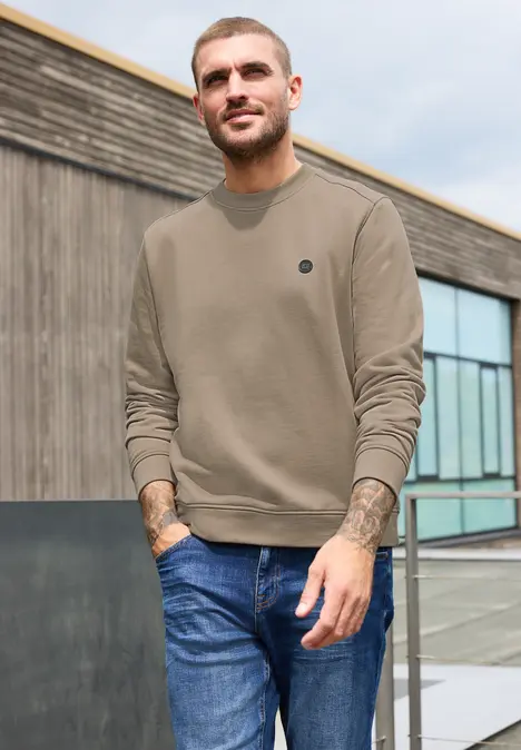 Ein lässiger Mann in olivfarbenem Pullover und blauen Jeans steht vor einem modernen Gebäude.
