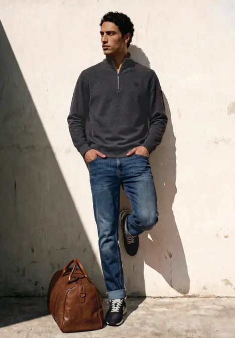 Ein Mann lehnt in lässigem Pullover und Jeans an einer Wand, neben ihm eine Leder-Reisetasche.