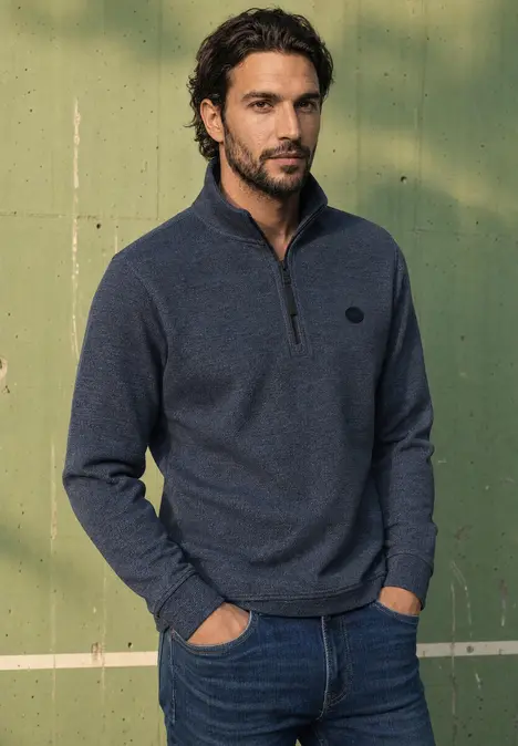 Ein junger Mann mit dunklem Bart trägt einen blauen Zip-Pullover und steht entspannt vor einer grünen Wand.