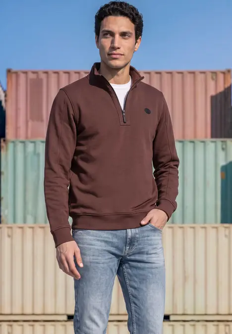 Ein Mann trägt braunen Half-Zip-Pullover und Jeans, steht vor bunten Containern.