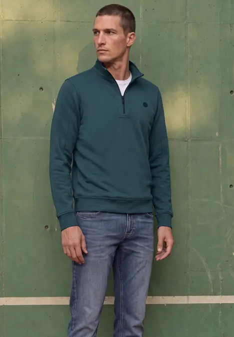 Ein Mann trägt einen dunkelgrünen Pullover mit halbem Reißverschluss und Jeans und steht vor einer grünen Wand.
