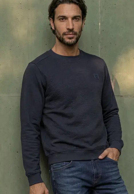 Ein Mann mit dunklem Sweatshirt und Jeans steht vor einer grünen Wand mit den Händen in den Taschen.