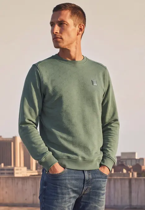 Een man staat buiten bij een hek, draagt een groen sweatshirt en spijkerbroek.