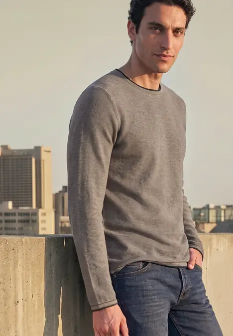 Mann in grauem Langarmshirt und Jeans lehnt auf einer Dachbarriere vor städtischer Skyline.