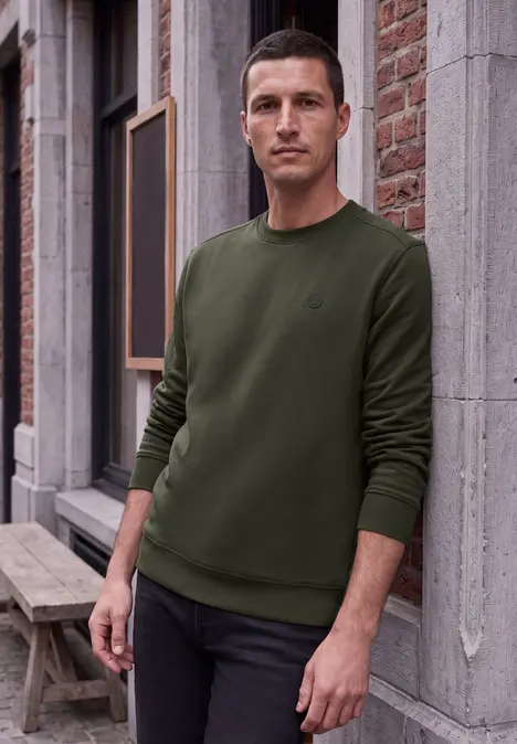 Ein Mann lehnt lässig an einer Ziegelwand, trägt grünen Pullover und schwarze Hose.