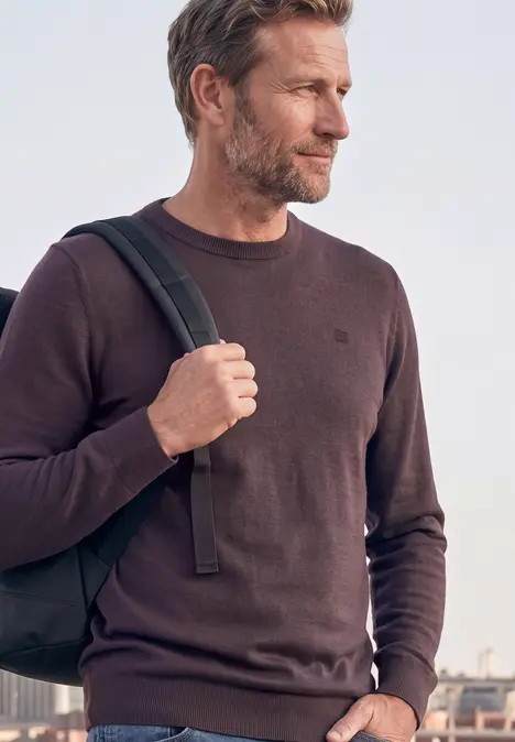 Ein Mann mittleren Alters trägt Rucksack und Pullover vor städtischer Kulisse.