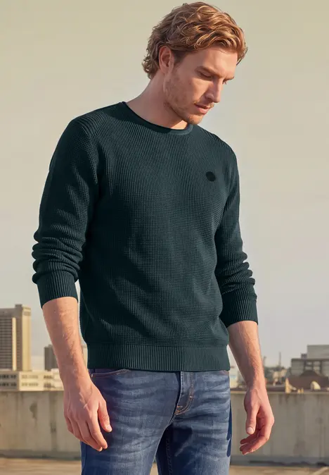 Un jeune homme se tient sur une terrasse en portant un pull noir et un jean bleu.