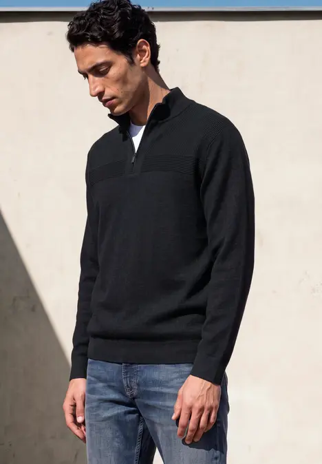 Ein Mann trägt einen schwarzen Pullover mit halbem Reißverschluss und blaue Jeans, er blickt nach unten.