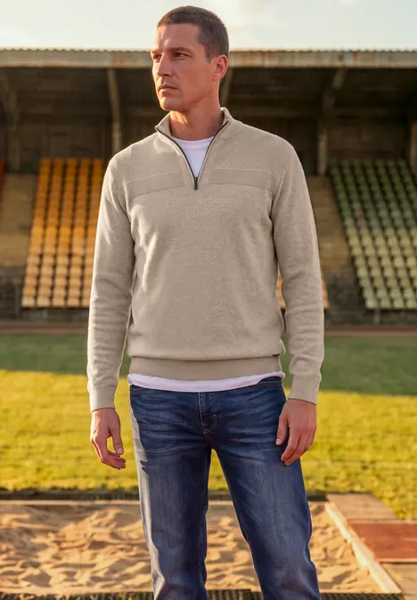 Ein Mann trägt Jeans und einen hellgrauen Pullover mit kurzem Reißverschluss auf einem Sportplatz.