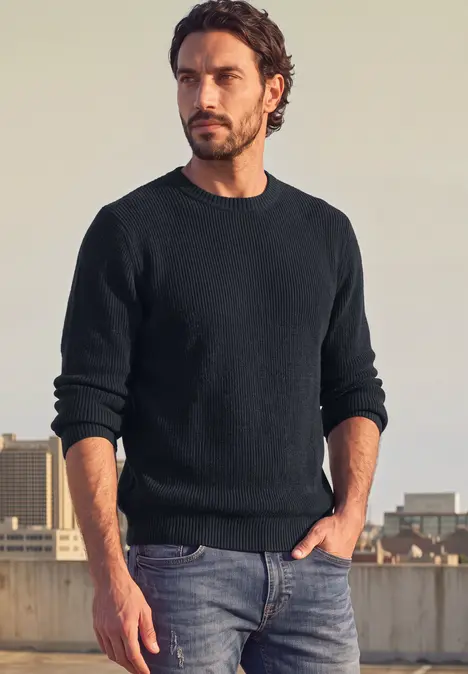 Ein Mann mit dunklem Pullover und Jeans auf einem Dach mit Stadtblick.