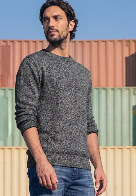 Ein Mann in grauem Pullover und Jeans steht vor farbigen Frachtcontainern