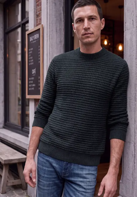 Ein Mann in grünem Pullover lehnt lässig an einer Hauswand vor einem Cafe