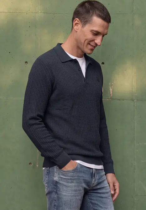 Mann im dunkelblauen Strick-Poloshirt mit weißem Kragen und Jeans vor grüner Wand, lächelnd.