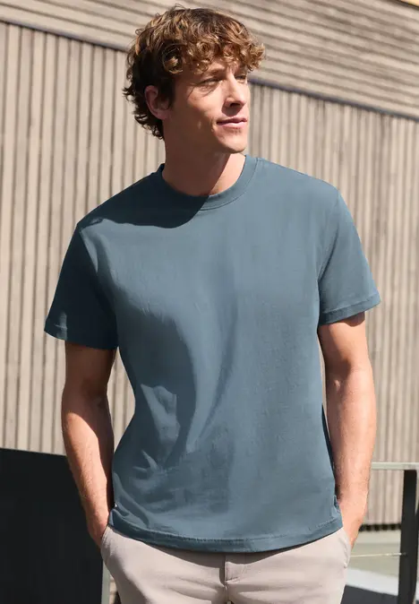 Ein junger Mann trägt ein blau-graues T-Shirt und steht mit den Händen in den Taschen vor einer Holzwand.