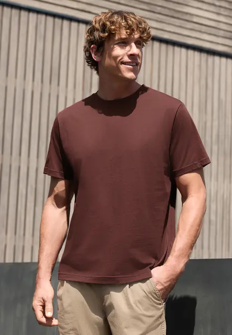 Ein Mann mit braunem T-Shirt und beigen Hosen steht vor einer Holzfassade, die Hände in den Taschen.