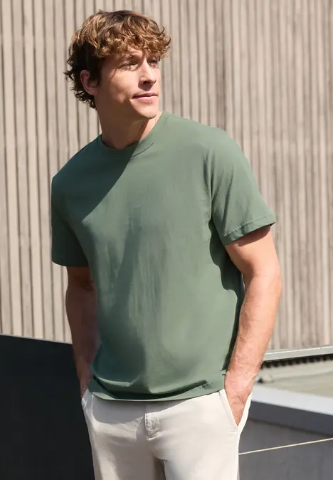 Ein Mann mit lockigem Haar trägt ein grünes T-Shirt und steht entspannt vor einer Holzfassade.