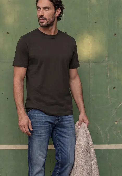 Mann vor grünem Hintergrund trägt dunkles T-Shirt, Jeans und hält eine beige Jacke.