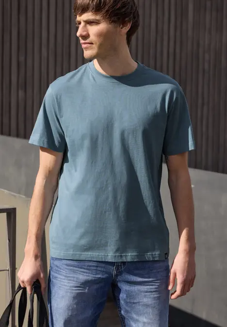 Ein Mann steht vor einer Holzwand, trägt ein blaues T-Shirt, Jeans und hält eine Tasche in der Hand.