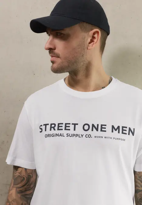 Mann mit Basecap und weißem STREET ONE MEN T-Shirt, tätowierte Arme, vor hellem Hintergrund.