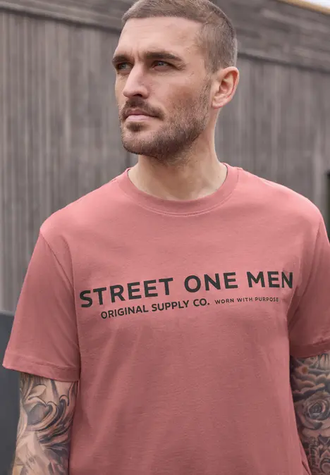 Ein tätowierter Mann trägt ein rosa T-Shirt mit dem Aufdruck 'STREET ONE MEN Original Supply Co.' vor einem Holzzaun.
