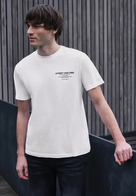 Ein Mann in weißem T-Shirt und dunkler Jeans lehnt an einer Mauer vor einer Holzfassade.