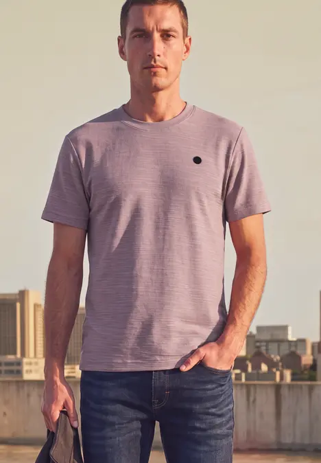 Un homme en jean et t-shirt se tient devant un décor urbain, tenant une petite sacoche avec décontraction.