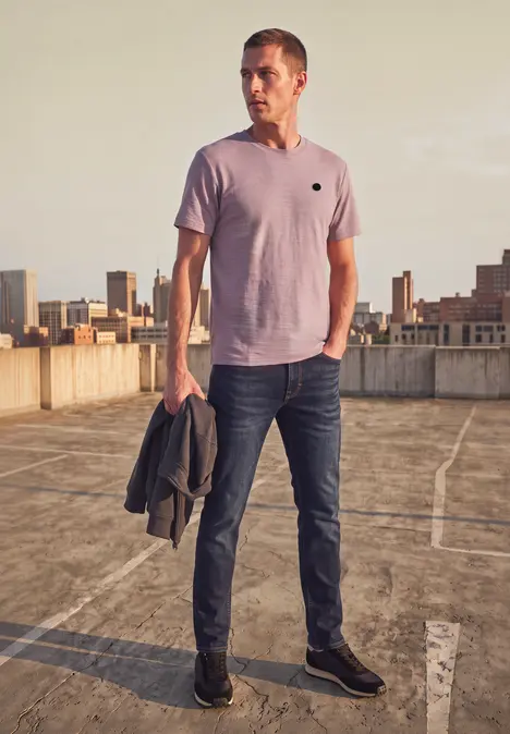 Ein Mann in Jeans und T-Shirt steht auf einem Parkdeck, hält eine Jacke, Stadtkulisse im Hintergrund.