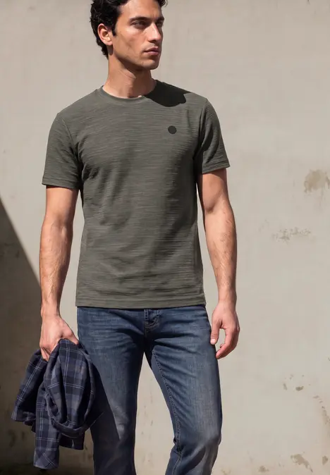 Een man draagt een kaki T-shirt en spijkerbroek en houdt een geruite blouse vast voor een neutrale achtergrond.
