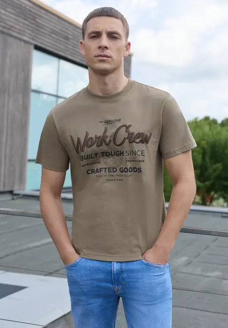 Ein junger Mann steht auf einer Dachterrasse, trägt ein khakifarbenes T-Shirt mit Aufdruck und eine blaue Jeans.
