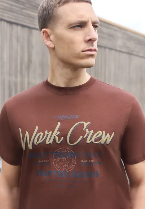 Ein Mann in braunem T-Shirt mit 'Work Crew'-Schriftzug und urbaner Holzwand im Hintergrund.