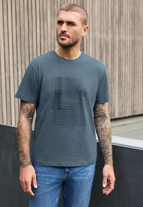 Een getatoeëerde man in T-shirt en spijkerbroek staat voor een houten wand en kijkt opzij.