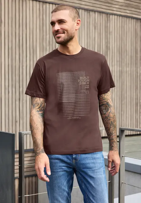 Ein lächelnder Mann mit braunem T-Shirt und Jeans steht vor Holzwand, trägt Tattoos.