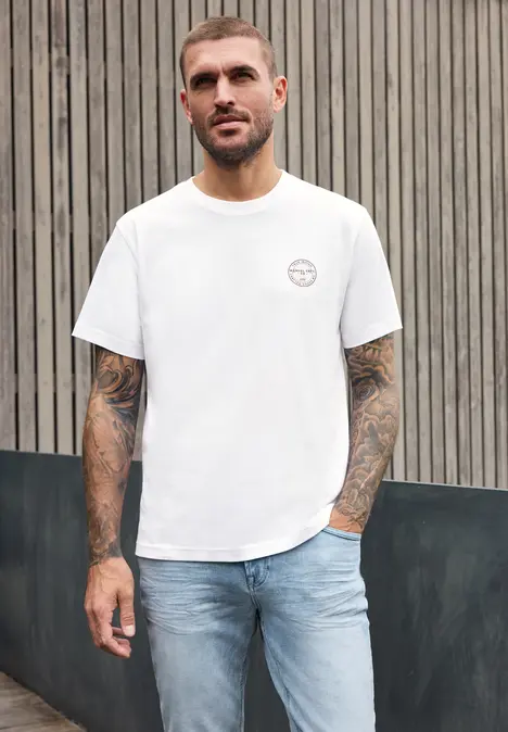 Ein tätowierter Mann trägt ein weißes T-Shirt und Jeans und steht vor einer hölzernen Wand.