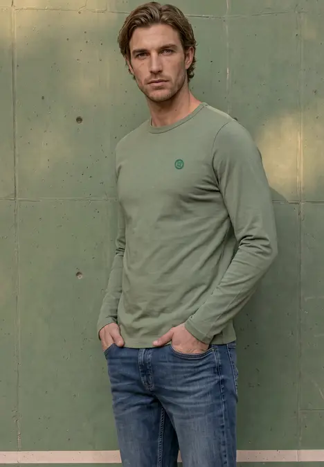 Ein junger Mann mit hellbraunen Haaren trägt ein grünes Langarmshirt und Jeans vor einer grünen Wand.