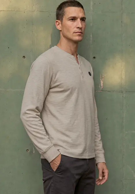 Ein Mann trägt ein graues Langarm-Henley-Shirt, steht seitlich mit Händen in den Hosentaschen vor einer grünen Wand.