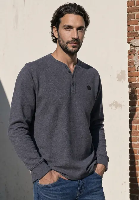 Ein Mann lehnt an einer Mauer und trägt ein dunkelblaues Henley-Shirt.