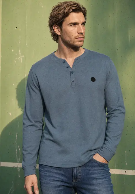 Ein Mann steht vor einer grünen Wand, trägt ein blaues, langärmeliges Shirt mit Knopfleiste am Kragen.