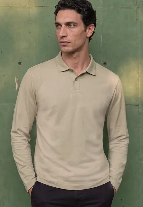 Ein Mann in khakifarbener Langarm-Poloshirt steht vor grünem Hintergrund und hat die Hände in den Taschen.