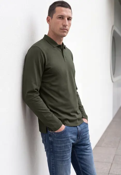 Ein Mann lehnt lässig in einem weißen Flur an einer Wand, trägt olivgrünes Langarm-Poloshirt und blaue Jeans.