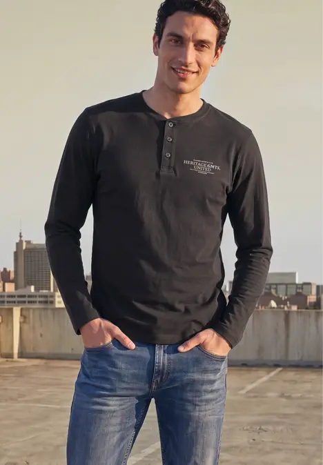 Ein Mann in dunklem Langarmshirt und blauen Jeans steht lächelnd auf einem Dach mit Stadthintergrund.