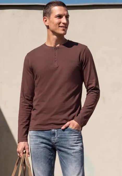Ein junger Mann trägt ein braunes Henley-Shirt und Jeans, hält eine geflochtene Tasche vor einer hellen Wand.