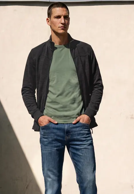Ein Mann in grünem T-Shirt, schwarzer Jacke und Jeans steht entspannt vor einer hellen Wand im Sonnenlicht.