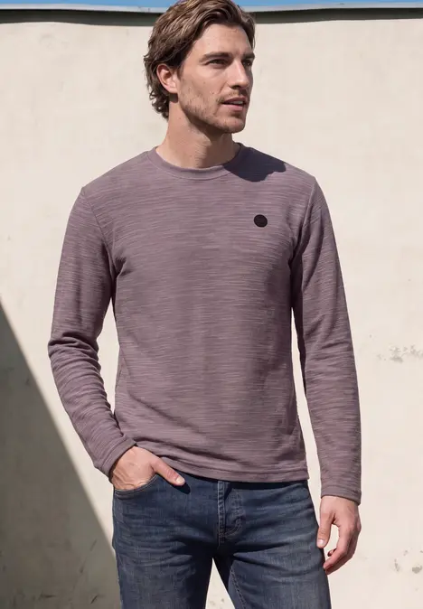 Ein Mann trägt ein lila Langarmshirt und Jeans, steht vor einer Wand im Sonnenlicht.