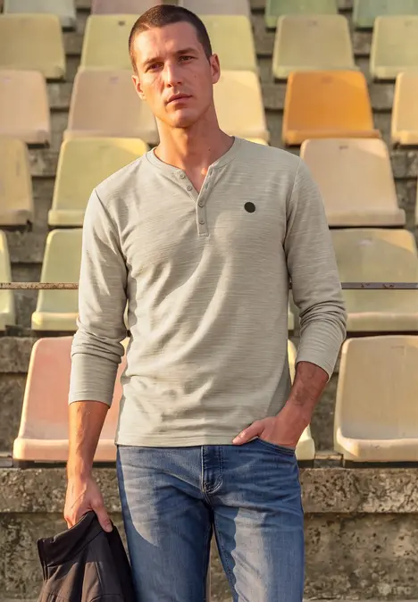Ein Mann steht vor leeren Stadionrängen, trägt ein graues Langarmshirt, blaue Jeans und hält eine Jacke.