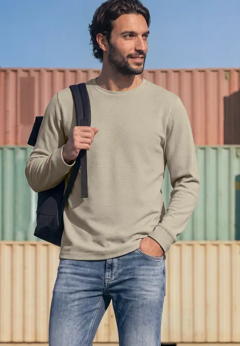 Mann in olivgrünem Pullover, Jeans und Rucksack vor bunten Frachtcontainern.