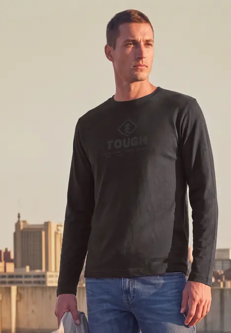 Un homme porte un sweat-shirt noir marqué 'TOUGH' sur un toit, avec une silhouette urbaine en arrière-plan.