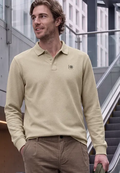 Mann in beigem Langarm-Poloshirt und brauner Hose, lässig stehend vor moderner Treppe.