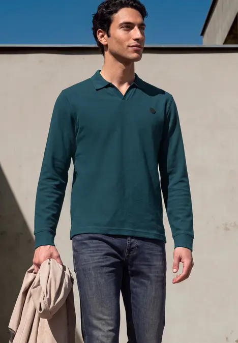 Ein Mann trägt ein petrolfarbenes Langarm-Poloshirt und dunkle Jeans und hält eine Jacke in der Hand.