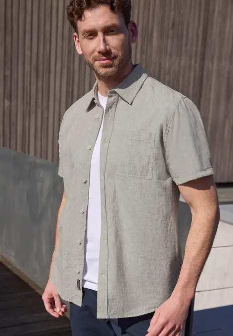 Un homme devant un mur en bois, portant une chemise claire sur un t-shirt blanc.