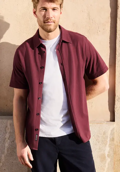 Un homme porte une surchemise bordeaux sur un T-shirt blanc et un pantalon sombre devant un mur beige.